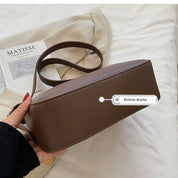 Borsa a tracolla marrone in pelle, design minimalista, con etichetta "Bottom display". Sfondo con rivista d'arte e tessuto beige.