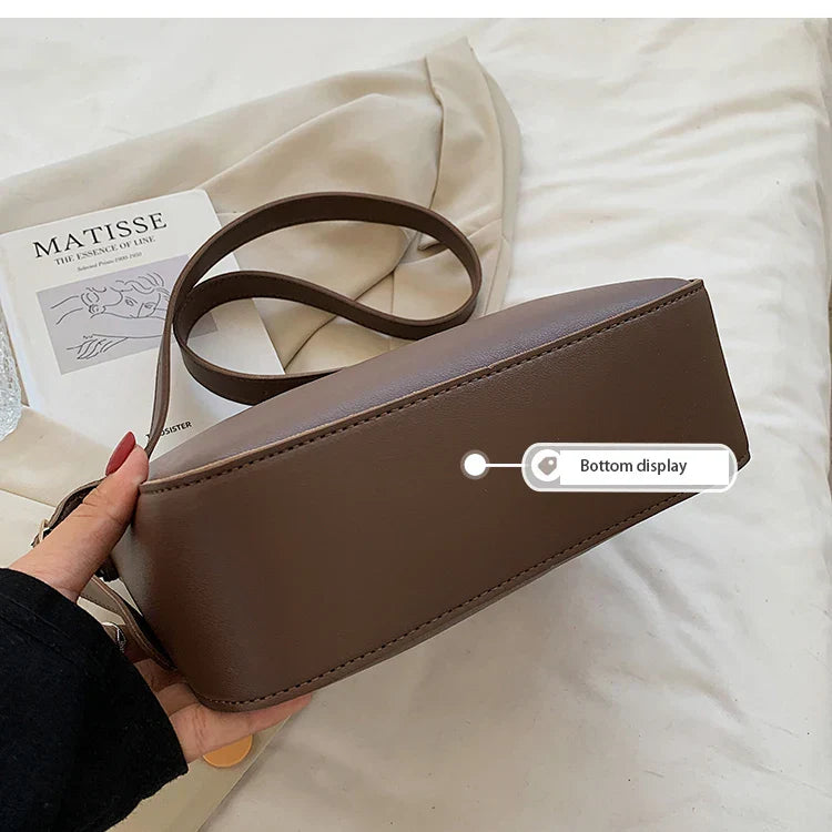 Borsa a tracolla marrone in pelle, design minimalista, con etichetta "Bottom display". Sfondo con rivista d'arte e tessuto beige.