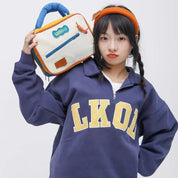 Ragazza con felpa blu "LKOL" e borsa colorata a mano, posa su sfondo bianco. Moda casual, accessori vivaci, stile giovanile.