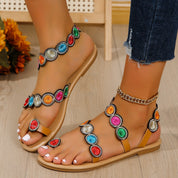 Sandali estivi da donna con cinturini decorati da pietre colorate, suola piatta beige, indossati con jeans. Accessorio moda casual e vivace.