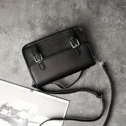Borsa a tracolla nera in pelle su sfondo grigio, con fibbie decorative e tracolla regolabile. Elegante accessorio moda per donna.