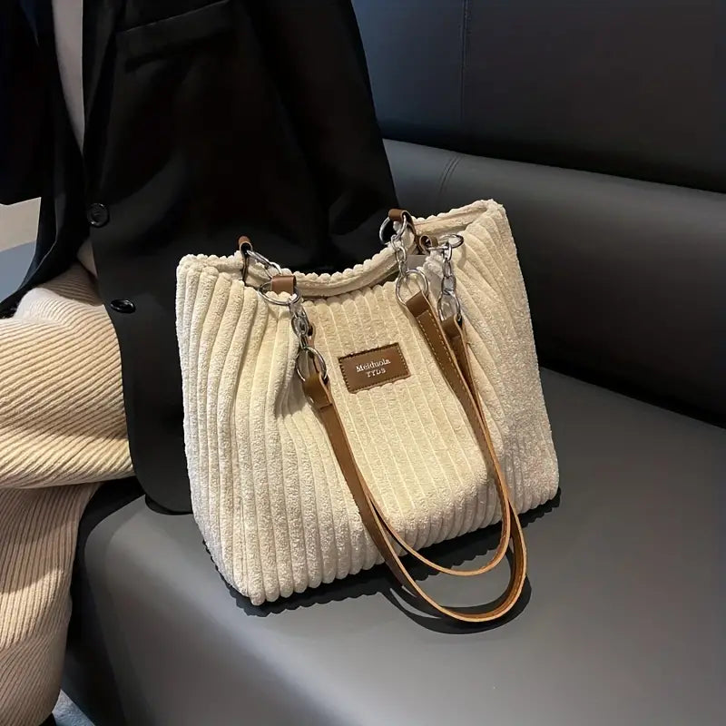 Borsa a spalla in tessuto a coste beige con manici in pelle marrone, dettagli metallici, su divano grigio. Elegante accessorio moda donna.
