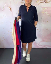 Donna con abito blu scuro, maniche a tre quarti, tiene abiti colorati su grucce. Sfondo decorativo rosa e bianco. Scarpe da ginnastica bianche.