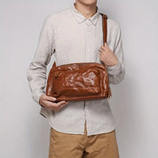 Uomo con camicia beige e pantaloni marroni tiene una borsa a tracolla in pelle marrone. Accessorio moda maschile, stile casual ed elegante.