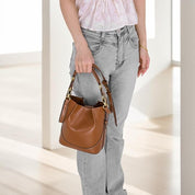 Donna con borsa a secchiello in pelle marrone, dettagli dorati, indossa jeans grigi e top bianco. Accessorio moda elegante e versatile.