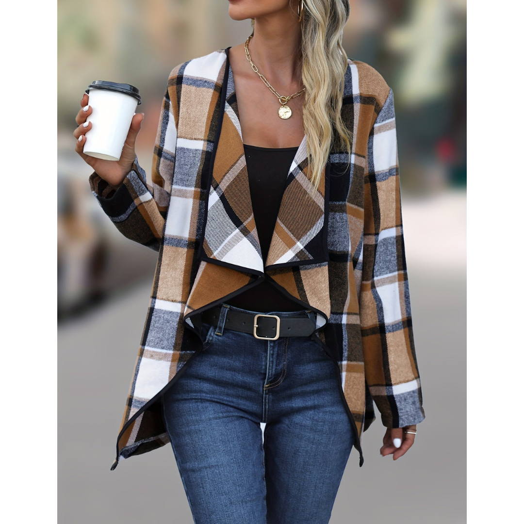 Donna con giacca a quadri marrone e bianca, top nero, jeans blu, cintura nera, collana dorata, tiene tazza da caffè. Moda autunnale casual.