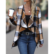 Donna con giacca a quadri marrone e bianca, top nero, jeans blu, cintura nera, collana dorata, tiene tazza da caffè. Moda autunnale casual.