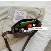 Borsa a tracolla marrone aperta su letto, contenente smartphone verde, rossetto, profumo, libro "Matisse", bicchiere e cappotto beige.