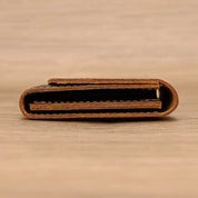 Portafoglio in pelle marrone chiaro, design minimalista, chiuso e visto di lato su sfondo in legno. Accessorio elegante e compatto.