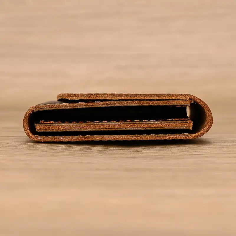 Portafoglio in pelle marrone chiaro, design minimalista, chiuso e visto di lato su sfondo in legno. Accessorio elegante e compatto.