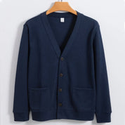 Cardigan blu scuro in cotone, con scollo a V, tre bottoni e due tasche frontali, appeso su gruccia di legno. Maglione elegante e casual.
