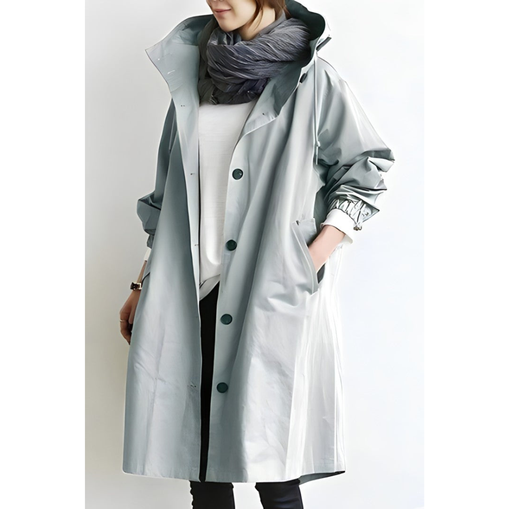 Cappotto impermeabile grigio chiaro da donna, lungo, con bottoni neri e cappuccio, indossato sopra maglia bianca e sciarpa grigia.