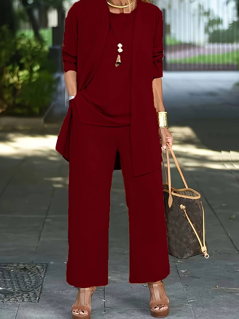 Donna in elegante completo rosso bordeaux, pantaloni larghi e giacca lunga, con borsa a tracolla marrone e sandali con tacco. Moda autunnale chic.