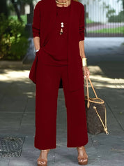 Donna in elegante completo rosso bordeaux, pantaloni larghi e giacca lunga, con borsa a tracolla marrone e sandali con tacco. Moda autunnale chic.