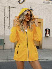 Giacca impermeabile gialla da donna con cappuccio, indossata da una modella all'aperto. Moda autunnale, stile casual, occhiali da sole.