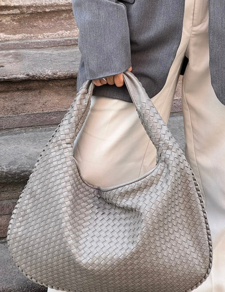 Borsa a spalla intrecciata in pelle grigia, indossata da una persona con giacca grigia e pantaloni bianchi. Accessorio moda elegante e moderno.
