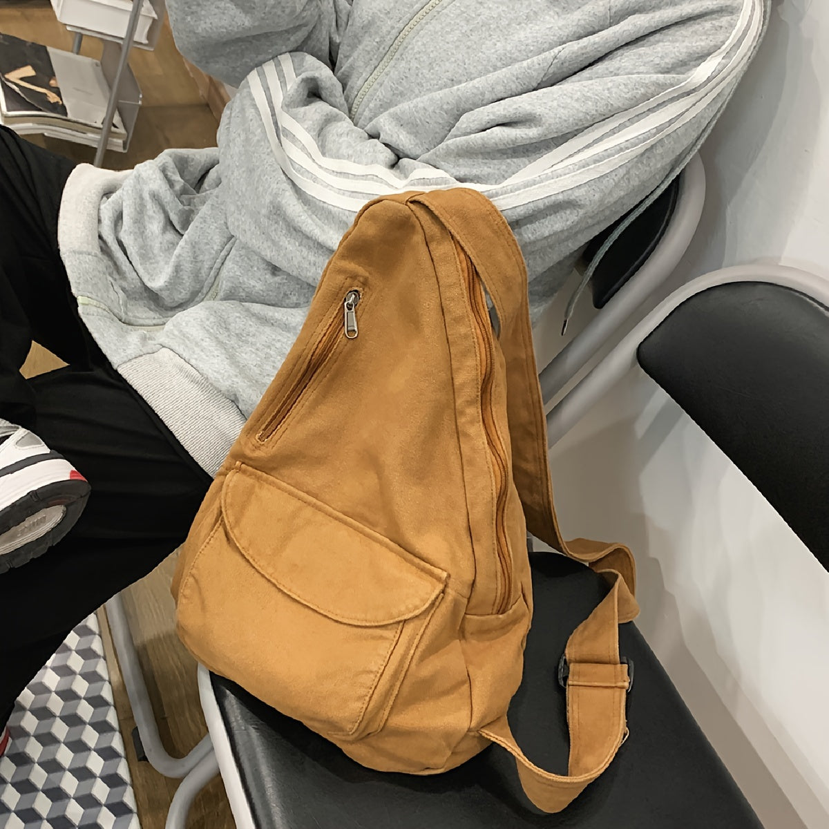 Zaino a tracolla in tessuto marrone su sedia, accanto a persona in felpa grigia. Design casual, ideale per scuola o viaggio.