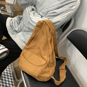 Zaino a tracolla in tessuto marrone su sedia, accanto a persona in felpa grigia. Design casual, ideale per scuola o viaggio.