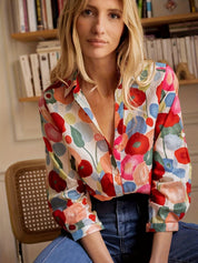 Donna bionda con camicia a fiori colorati, seduta su sedia di legno, sfondo con libreria. Moda femminile, abbigliamento casual, stile floreale.