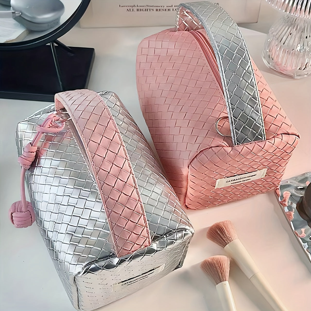 Due beauty case intrecciati, uno rosa e uno argento, su un tavolo con pennelli trucco. Eleganti e moderni, ideali per organizzare cosmetici.