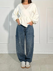 Donna con maglione bianco avvolgente e jeans larghi blu con effetto spruzzato, posa in interni. Look casual chic, moda autunno inverno 2023.