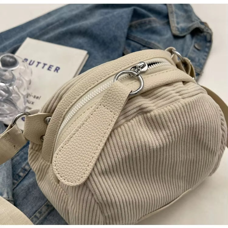 Borsa a tracolla beige in tessuto a coste, con zip argentata, su sfondo di jeans e libro. Accessorio moda casual, elegante e versatile.