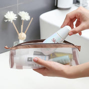 Mani che inseriscono cosmetici in una pochette trasparente da viaggio, bagno moderno sullo sfondo. Organizzatore trucco, accessori da viaggio.