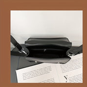 Borsa a tracolla nera in pelle, vista dall'alto, con interno spazioso e chiusura a zip, su sfondo di pagine di libro. Elegante e moderna.