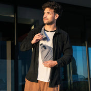Giovane uomo con marsupio grigio, camicia nera e pantaloni marroni, in piedi all'aperto. Accessorio moda uomo, stile casual urbano.