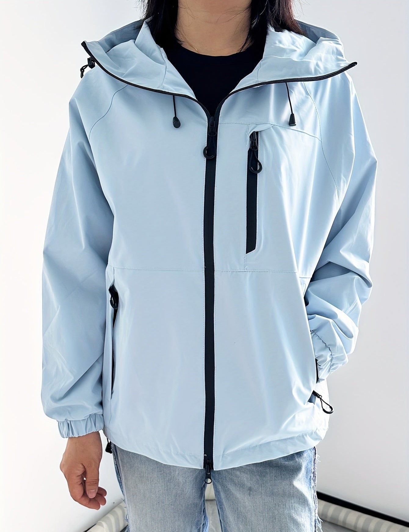 Giacca a vento unisex azzurra con cappuccio, zip frontale nera e tasche laterali, indossata da una persona. Abbigliamento casual, moda outdoor.