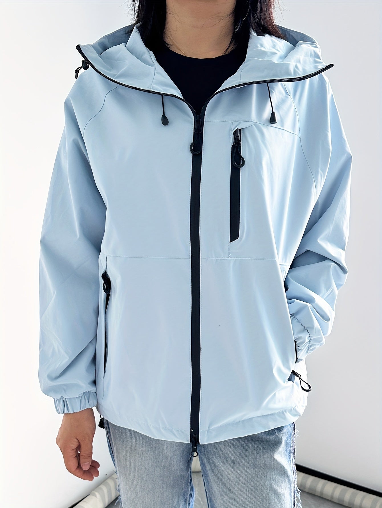Giacca a vento unisex azzurra con cappuccio, zip frontale nera e tasche laterali, indossata da una persona. Abbigliamento casual, moda outdoor.