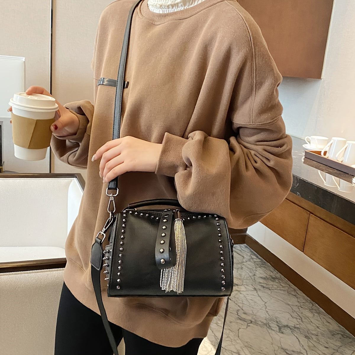 Donna con borsa a tracolla nera con borchie e frange, indossa maglione marrone, tiene bicchiere di caffè. Accessorio moda elegante e casual.