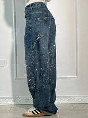 Jeans larghi in denim blu con dettagli scintillanti, tasche laterali, indossati con sneakers bianche e nere, sfondo interno neutro.