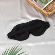 Maschera per dormire nera su tessuto bianco, con cinturino regolabile. Accessorio per il sonno, relax e viaggio. Sfondo con pianta e cesto.