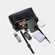 Borsa a tracolla nera in pelle con texture coccodrillo, contenente smartphone, profumo, chiavi, rossetto, auricolari e fazzoletti.