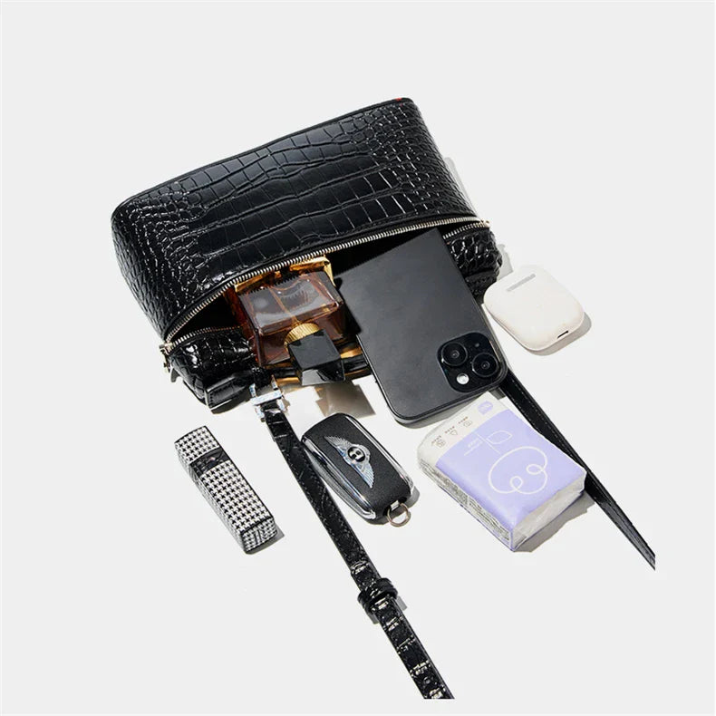 Borsa a tracolla nera in pelle con texture coccodrillo, contenente smartphone, profumo, chiavi, rossetto, auricolari e fazzoletti.
