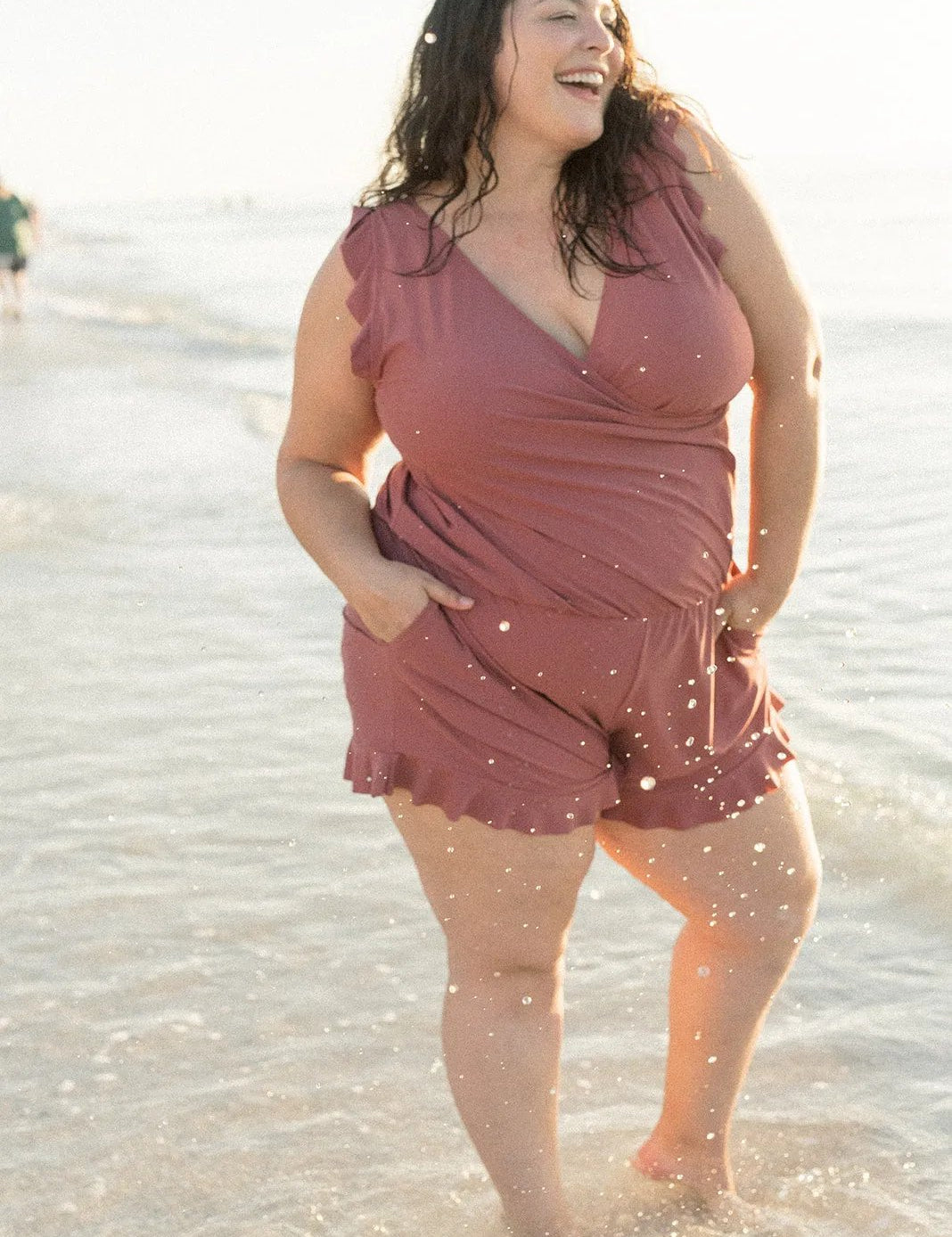 Donna sorridente in spiaggia con tuta rosa, moda mare taglie forti, estate, abbigliamento comodo, acqua, sole, felicità, stile casual.