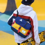 Giovane con felpa multicolore e zaino a tracolla blu e giallo, tiene uno skateboard. Moda urbana, accessori streetwear, stile casual.