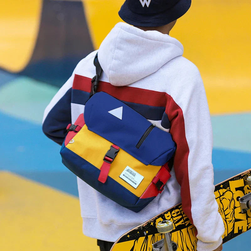 Giovane con felpa multicolore e zaino a tracolla blu e giallo, tiene uno skateboard. Moda urbana, accessori streetwear, stile casual.