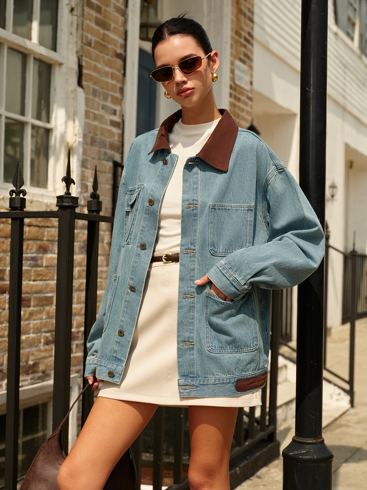 Donna con giacca di jeans oversize e abito bianco, occhiali da sole, borsa marrone, posa urbana. Moda street style autunnale.