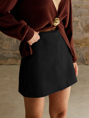 Gonna nera a vita alta, abbinata a maglione marrone con dettaglio dorato, moda autunno inverno, stile casual chic, sfondo muro in pietra.