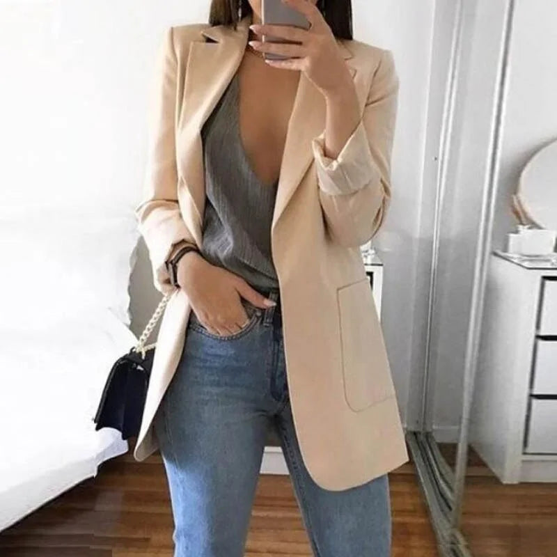 Donna con blazer beige elegante, top grigio scollato e jeans blu, in una stanza moderna. Moda casual chic, abbigliamento femminile trendy.