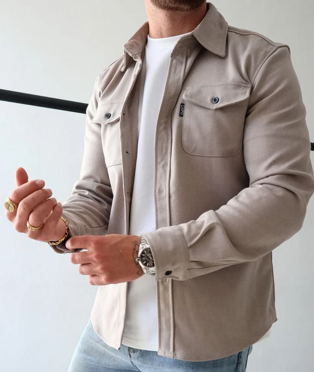Camicia beige da uomo in cotone, stile casual, con tasche frontali e bottoni, indossata sopra una maglietta bianca. Accessori: orologio e anelli.