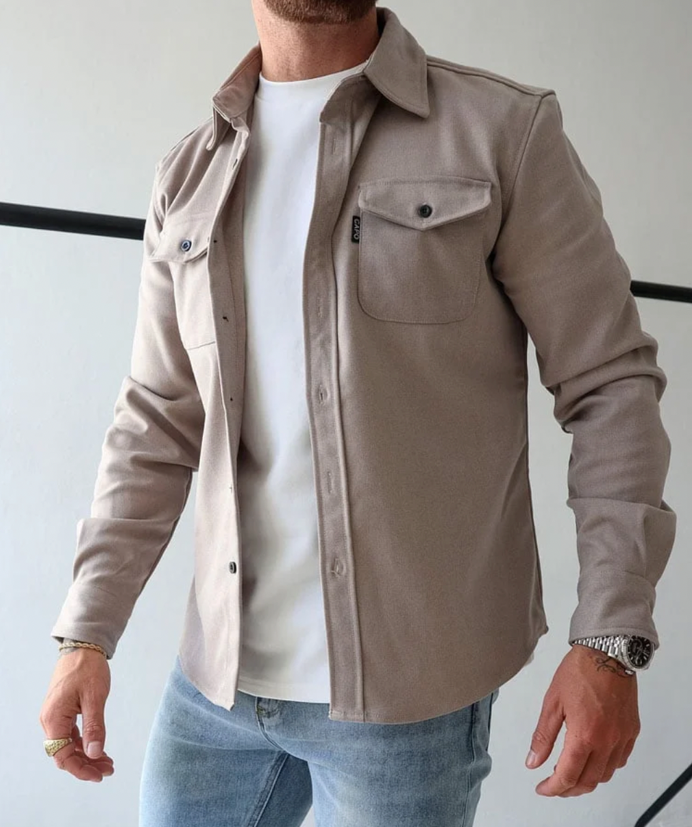 Camicia beige da uomo in cotone, stile casual, con tasche frontali, indossata sopra una maglietta bianca e jeans. Look moderno e versatile.