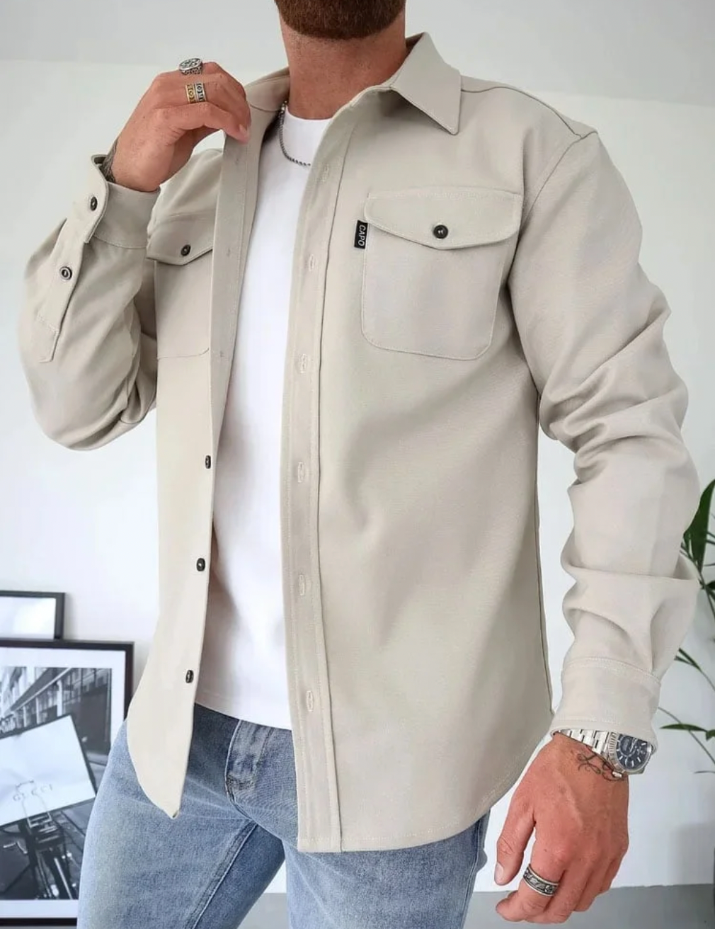 Camicia beige da uomo con tasche frontali, indossata sopra una maglietta bianca. Abbinata a jeans blu chiaro. Stile casual ed elegante.