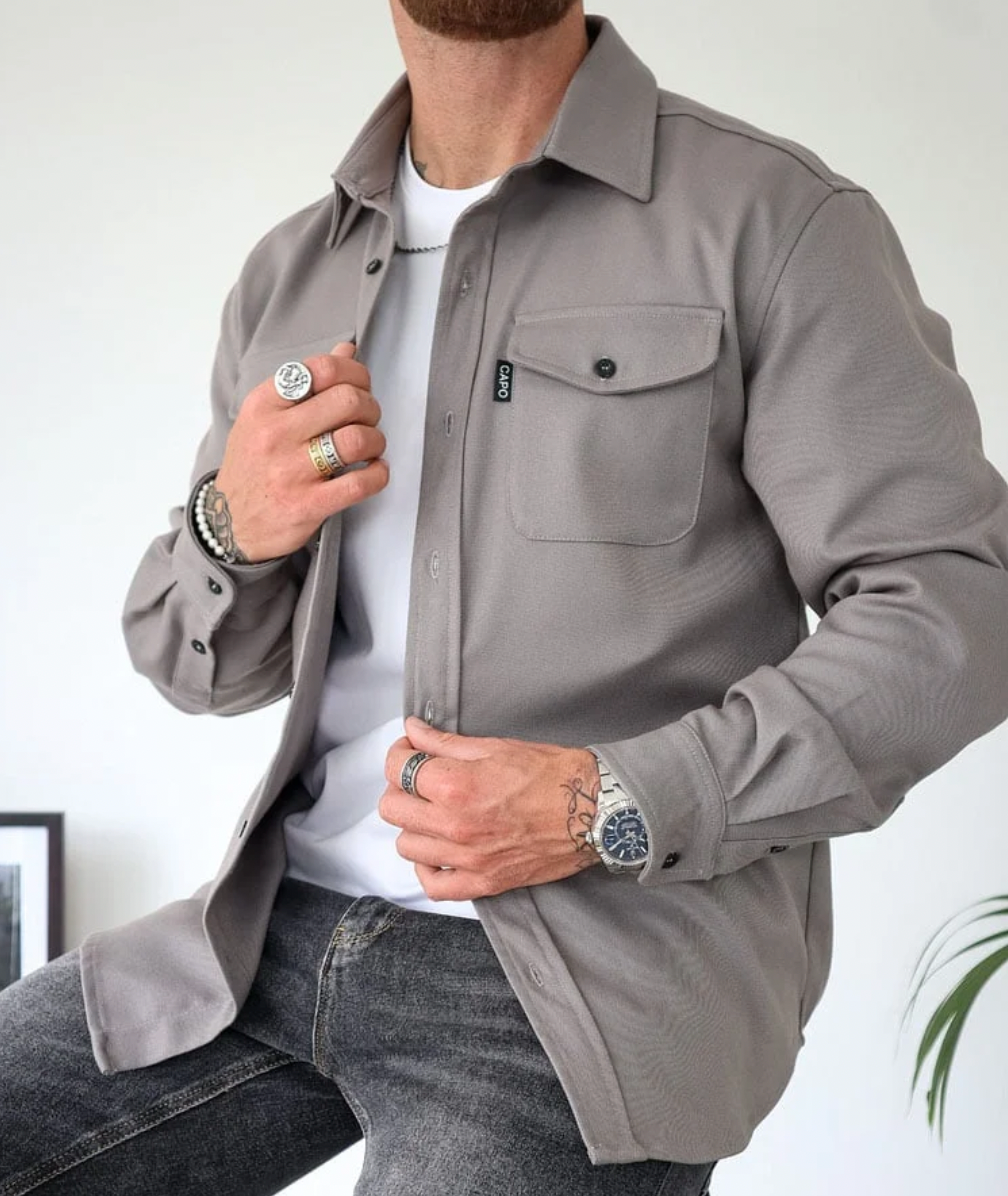 Camicia grigia da uomo, stile casual, con tasca frontale, indossata sopra una maglietta bianca. Accessori: orologio e anelli. Moda maschile elegante.