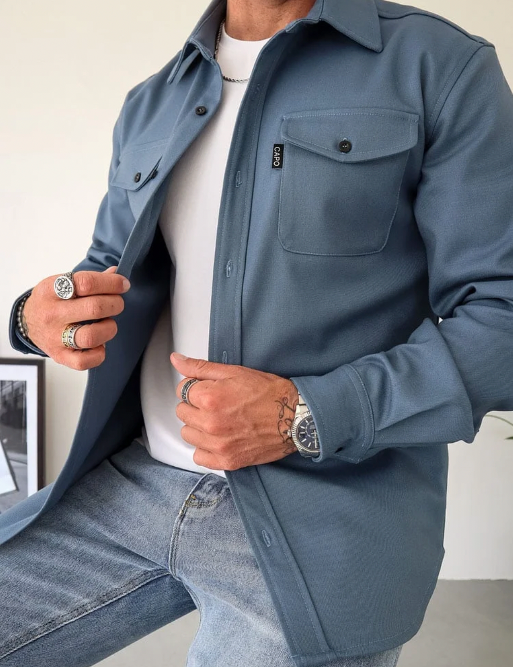 Camicia uomo blu scuro, stile casual, con tasche frontali e bottoni, indossata sopra t-shirt bianca e jeans. Accessori: anelli e orologio.
