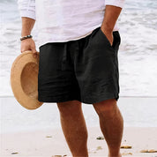 Uomo in spiaggia con pantaloncini neri e camicia bianca, tiene cappello di paglia. Moda estiva maschile, abbigliamento casual mare.