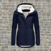 Giacca invernale donna blu con cappuccio foderato in pelliccia sintetica, chiusura a zip, tasche frontali, su sfondo muro di mattoni grigi.