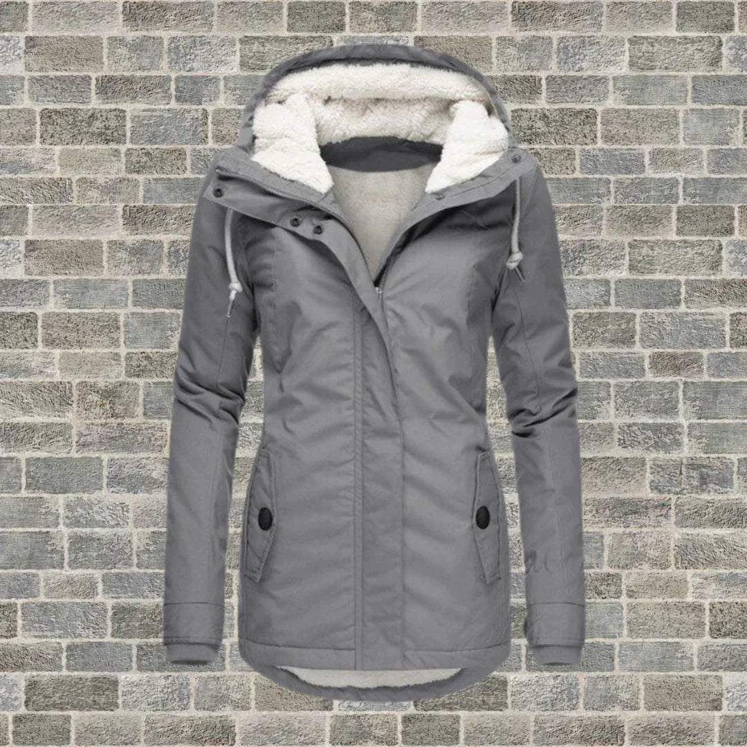 Giacca invernale da donna grigia con cappuccio foderato in pelliccia sintetica, chiusura a zip, tasche frontali, su sfondo di mattoni grigi.